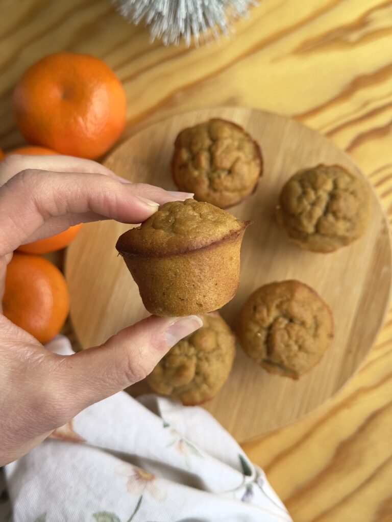 Mano sosteniendo un mini muffin de banana y mandarina sin azúcar añadida con más muffins al fondo