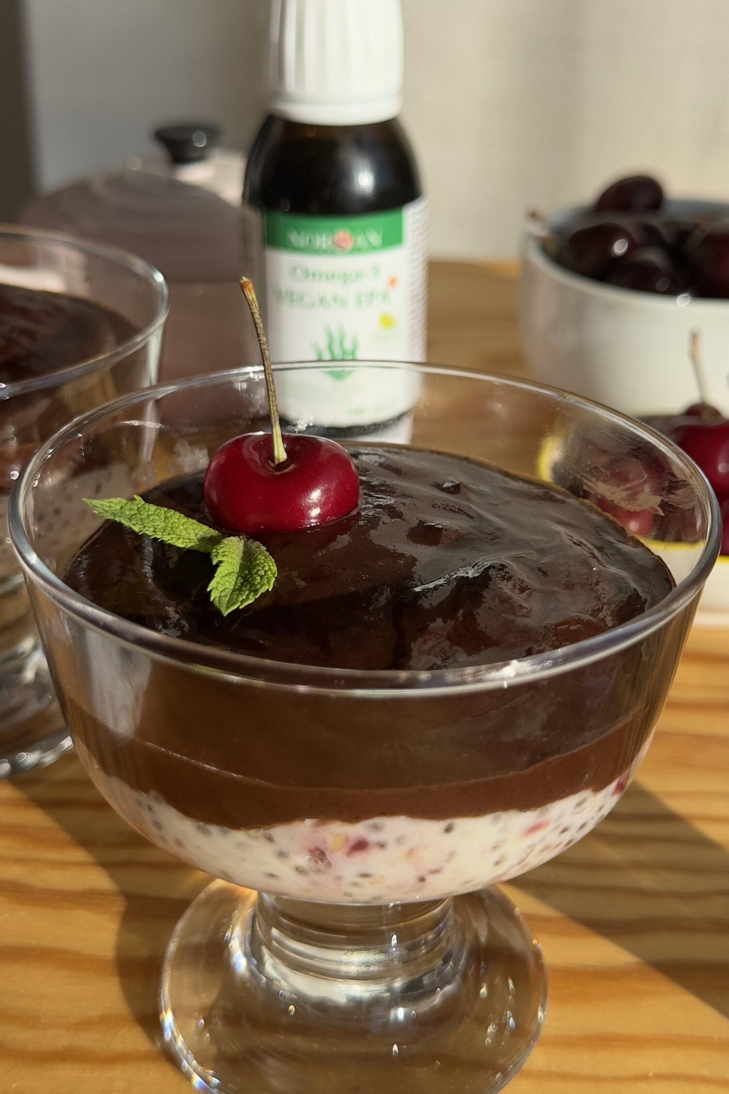Vasito de cristal con las capas del pudding de chía, mermelada de cerezas y ganache vegana de chocolate, decorado con cereza fresca y menta